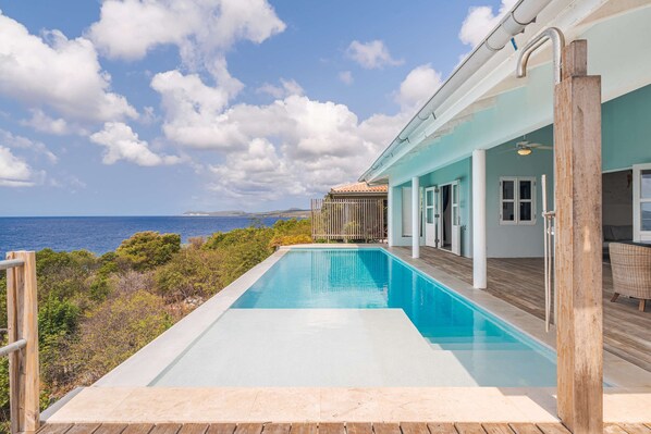 A pool with a wooden deck, a view of the sea, and a building with a light blue exterior. - Petite Oceanfront Retreat, 2 Bedrooms, Private Infinity Pool, Serene (Kralendijk)