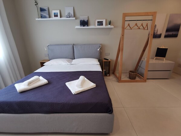 Un lit bien fait, avec des serviettes pliées, une table de chevet avec une télécommande, une petite armoire en bois et une étagère murale ornée de tableaux encadrés.