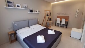 Ett modernt hotellrum med en stor säng, ett skrivbord och en stol. Det finns hyllor med bilder och ett litet bord med en lampa.