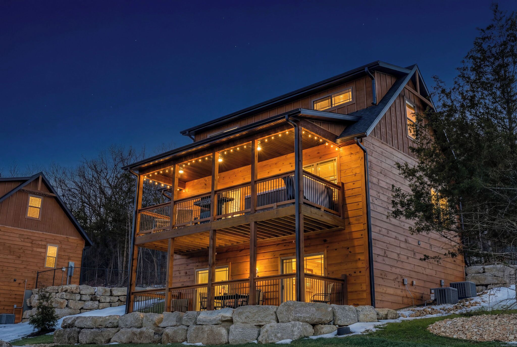 A two-story wooden cabin with a balcony, stone foundation, and string lights.