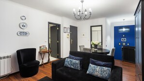 Un salon moderne avec un canapé noir, une petite table d’appoint et une table à manger avec des chaises. On y trouve également un fauteuil noir, un radiateur et un mur d’accent bleu.