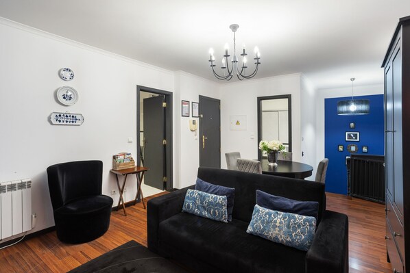 Un salon moderne avec un canapé noir, une petite table d’appoint et une table à manger avec des chaises. On y trouve également un fauteuil noir, un radiateur et un mur d’accent bleu.
