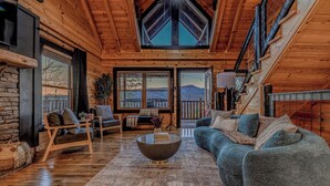 A cozy log cabin interior with a stone fireplace, a large window with a view, a wooden staircase, and a comfortable seating area. - *Spacious 5BD Lodge w/ VIEWS | Theater + Hot Tub* (Sevierville)