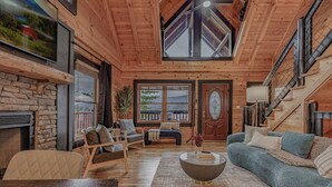 A cozy log cabin interior with a stone fireplace, wooden furniture, and a large window offering a view of the outdoors. - *Spacious 5BD Lodge w/ VIEWS | Theater + Hot Tub* (Sevierville)