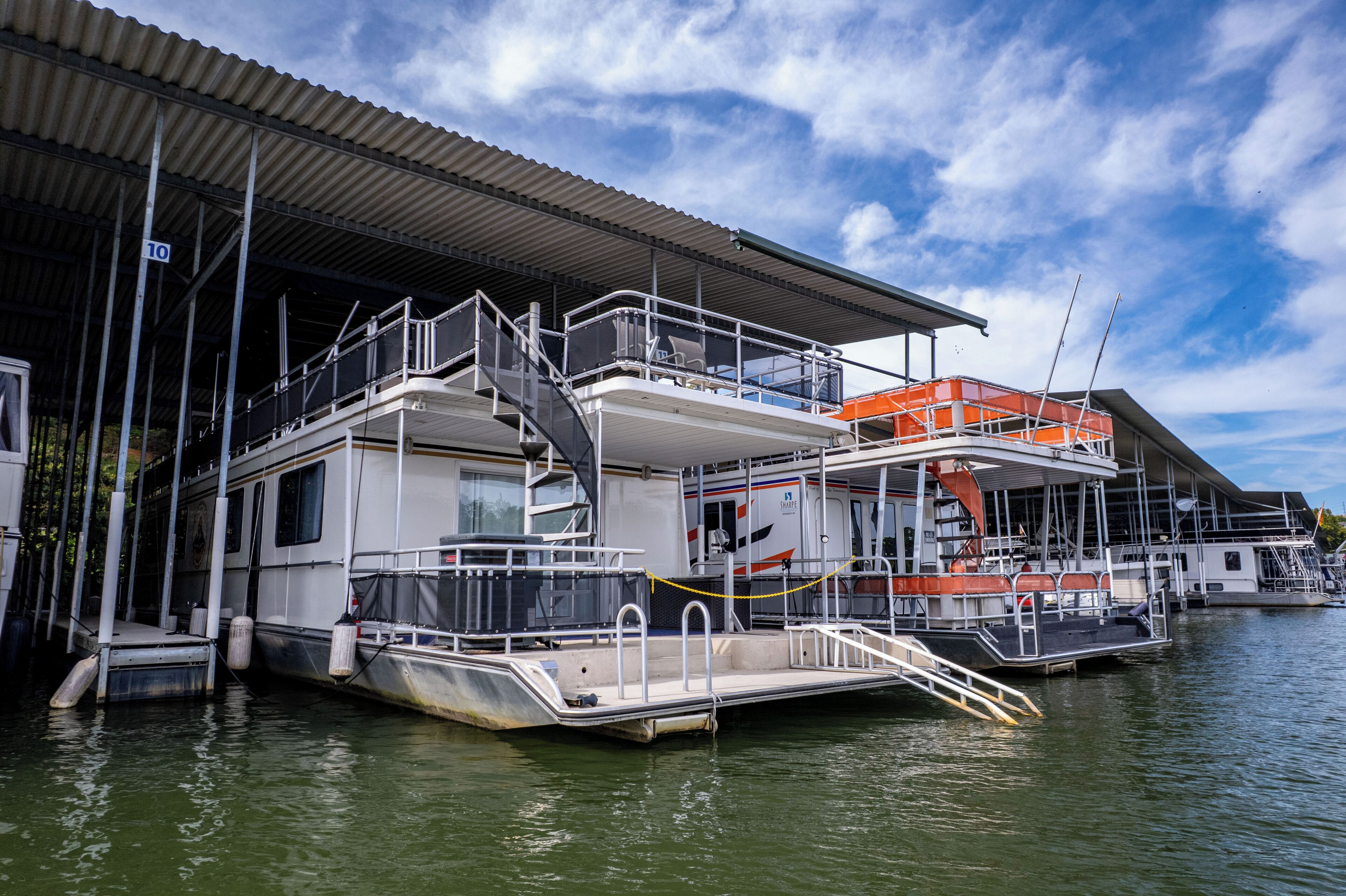 Une péniche amarrée dans une marina, dotée d’une terrasse couverte, d’un escalier et d’un canot de sauvetage.