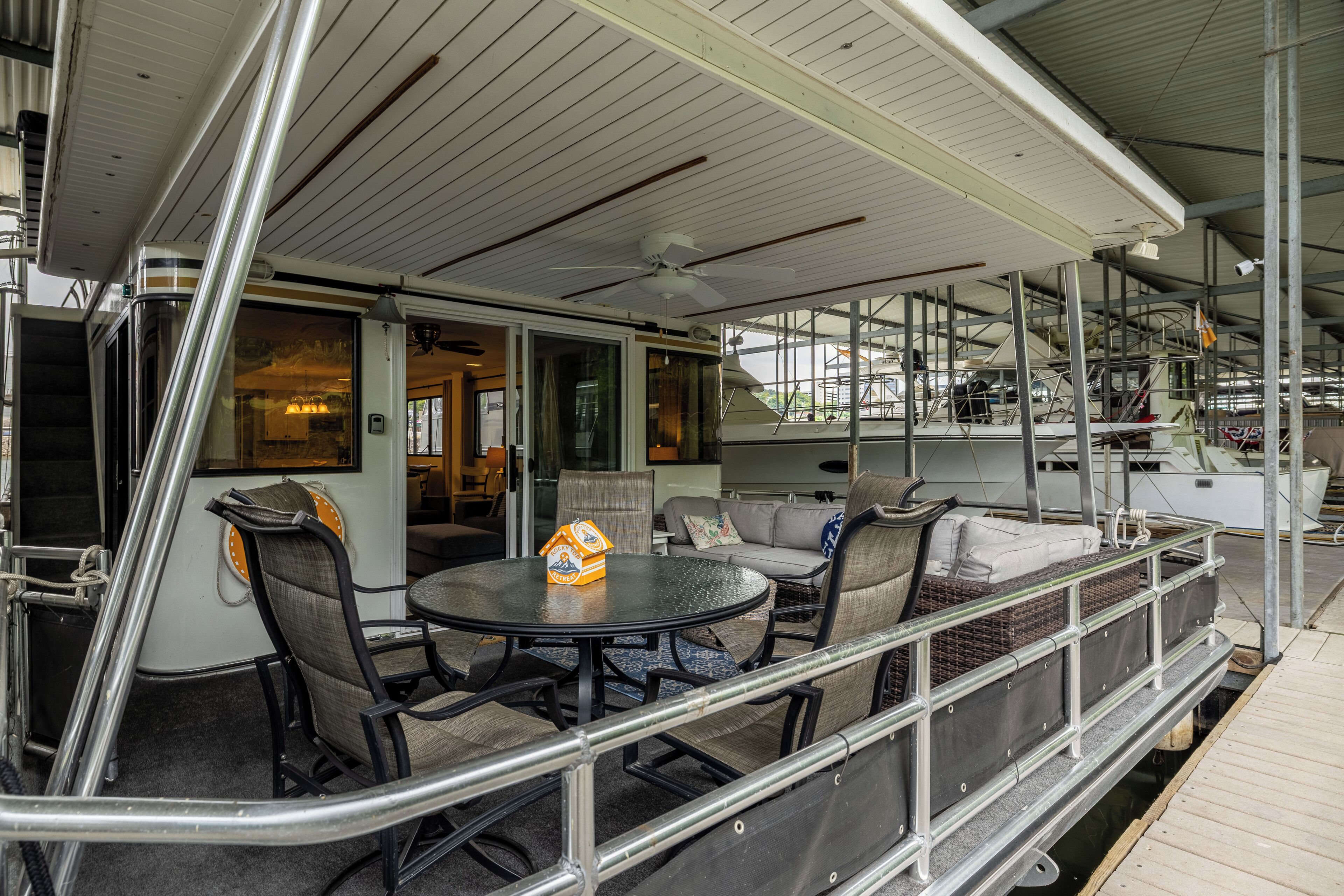 Une terrasse extérieure couverte, avec une table ronde et des chaises, une petite décoration en forme de maison et une vue sur l’eau.