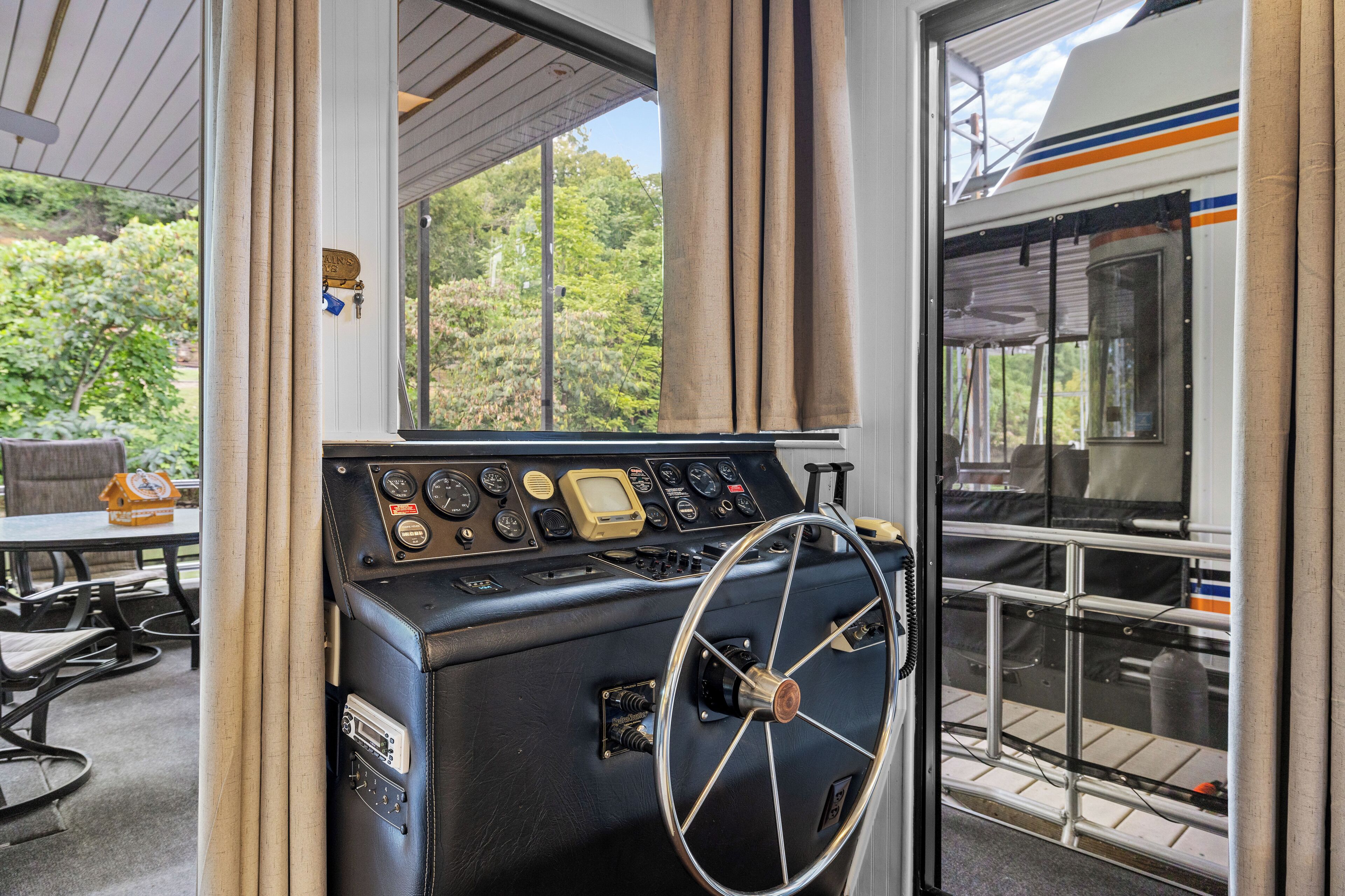 Un panel de control con indicadores y un volante, una ventana con cortinas, y vistas de vegetación al exterior.