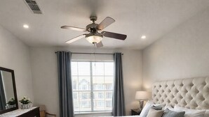 Ein Schlafzimmer mit einem großen Bett, einem Deckenventilator, einem Nachttisch, einer Kommode, einem Spiegel und einem Fenster mit Vorhängen.