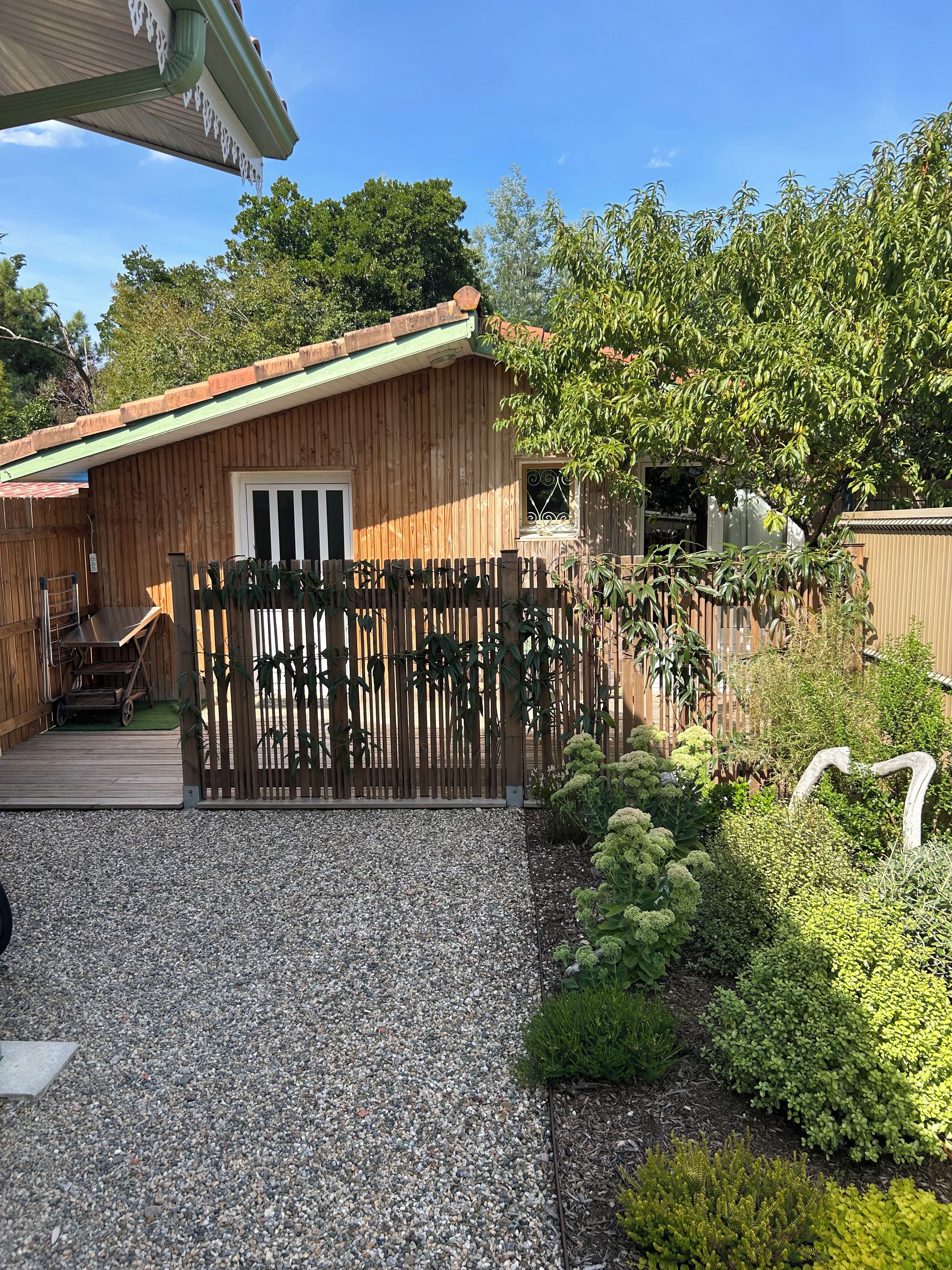 A wooden house with a gravel driveway, a wooden fence, and various plants.