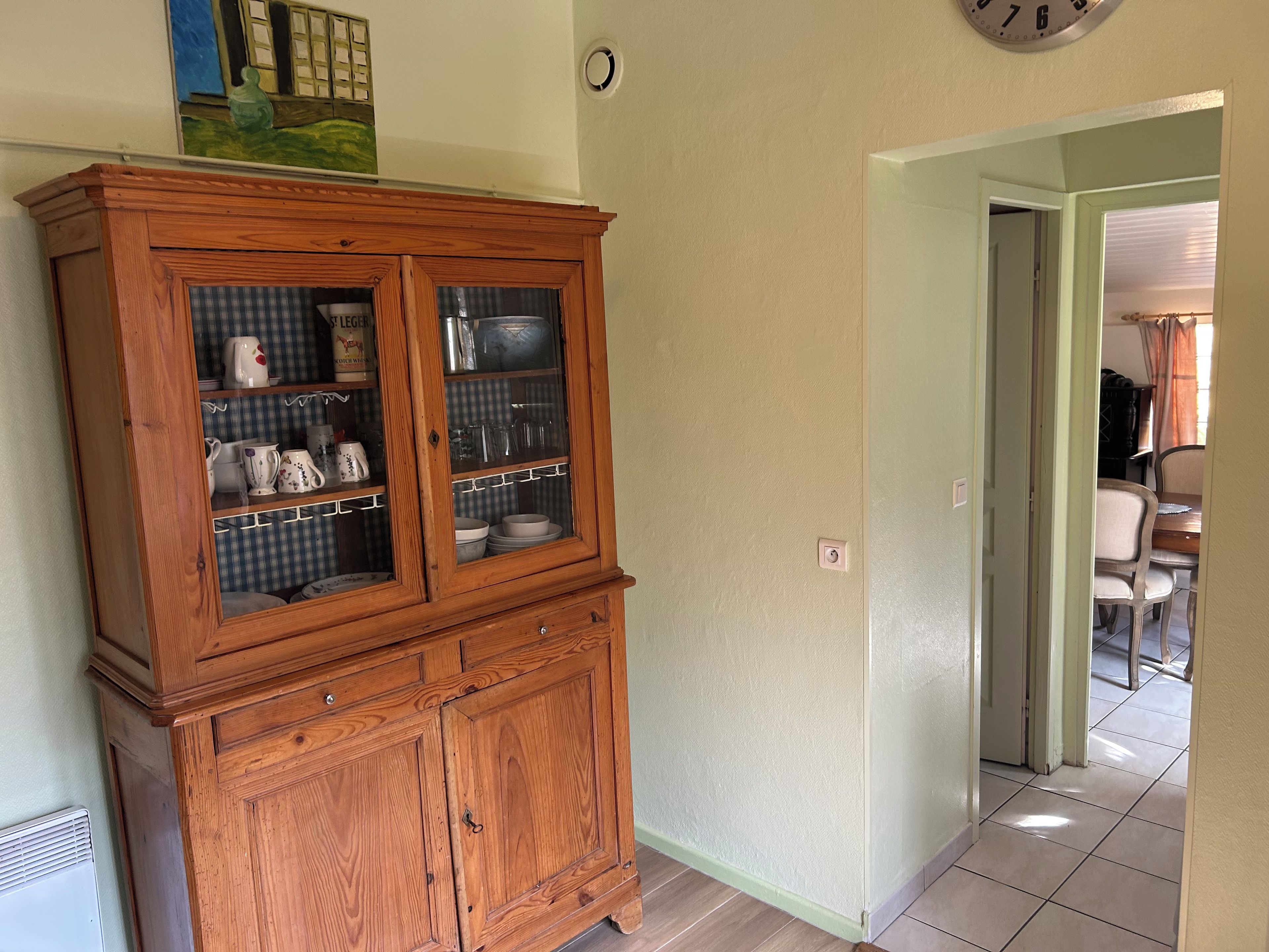 Un meuble en bois avec des portes en verre, une horloge au mur et un coin repas visible à travers une porte ouverte.