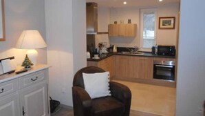 A modern kitchen with wooden cabinets, a microwave, and an oven. There is a dining area with a chair and a small table.