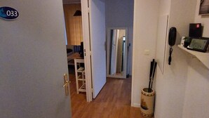 Un passadís amb sòl de fusta, una porta amb la inscripció '033', un petit cub de basures i una estant amb un telèfon i un objecte petit.