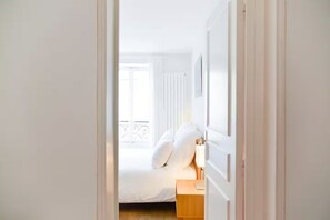 Un estrecho pasillo que conduce a un dormitorio con cama, mesita de noche y ventana con vistas al balcón.