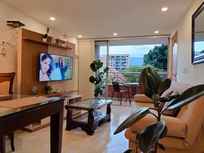 Ein modernes Wohnzimmer mit Fernseher, einem Glastisch, einer Couch und einem Balkon mit Blick auf Gebäude und Bäume.