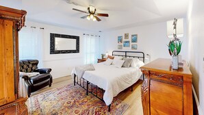 Ein Schlafzimmer mit einem Bett, einem Deckenventilator, einem Nachttisch, einer Kommode, einem Sessel, einem Teppich und einem Spiegel an der Wand.
