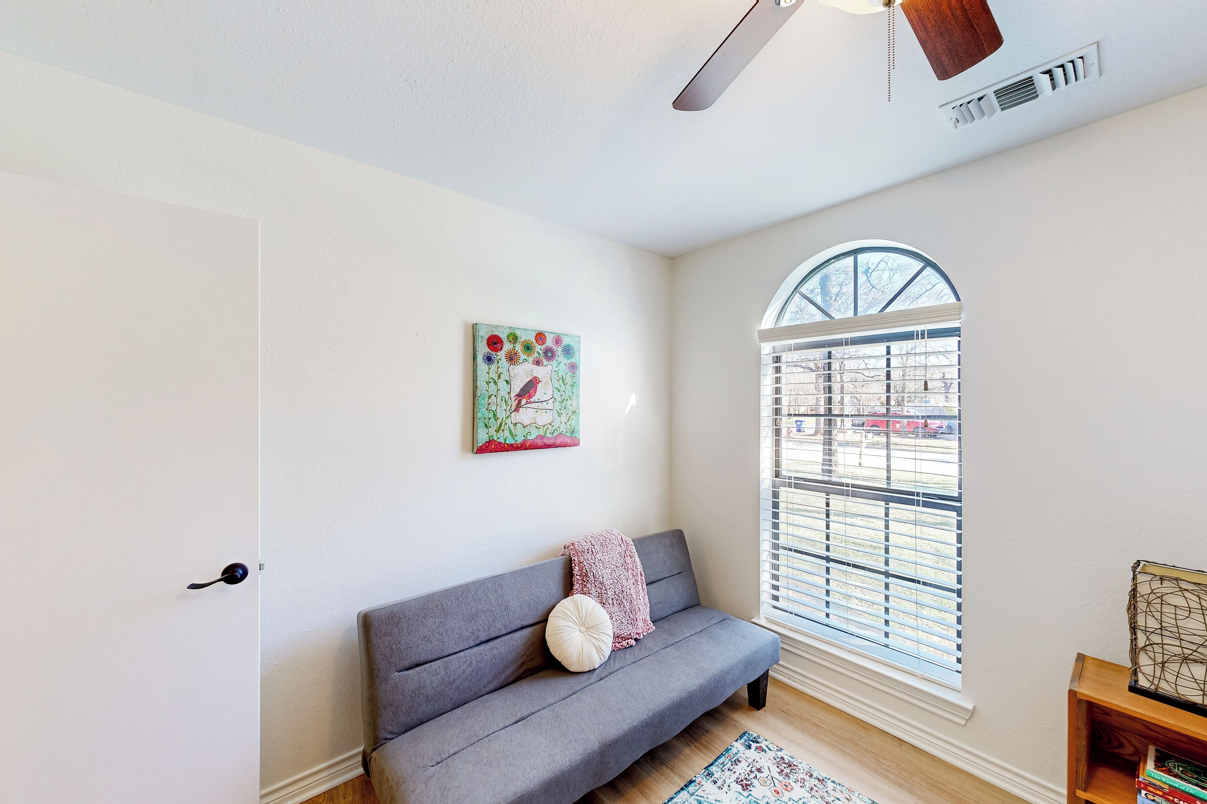 A room with a grey sofa, a window with blinds, a ceiling fan, and a painting on the wall.
