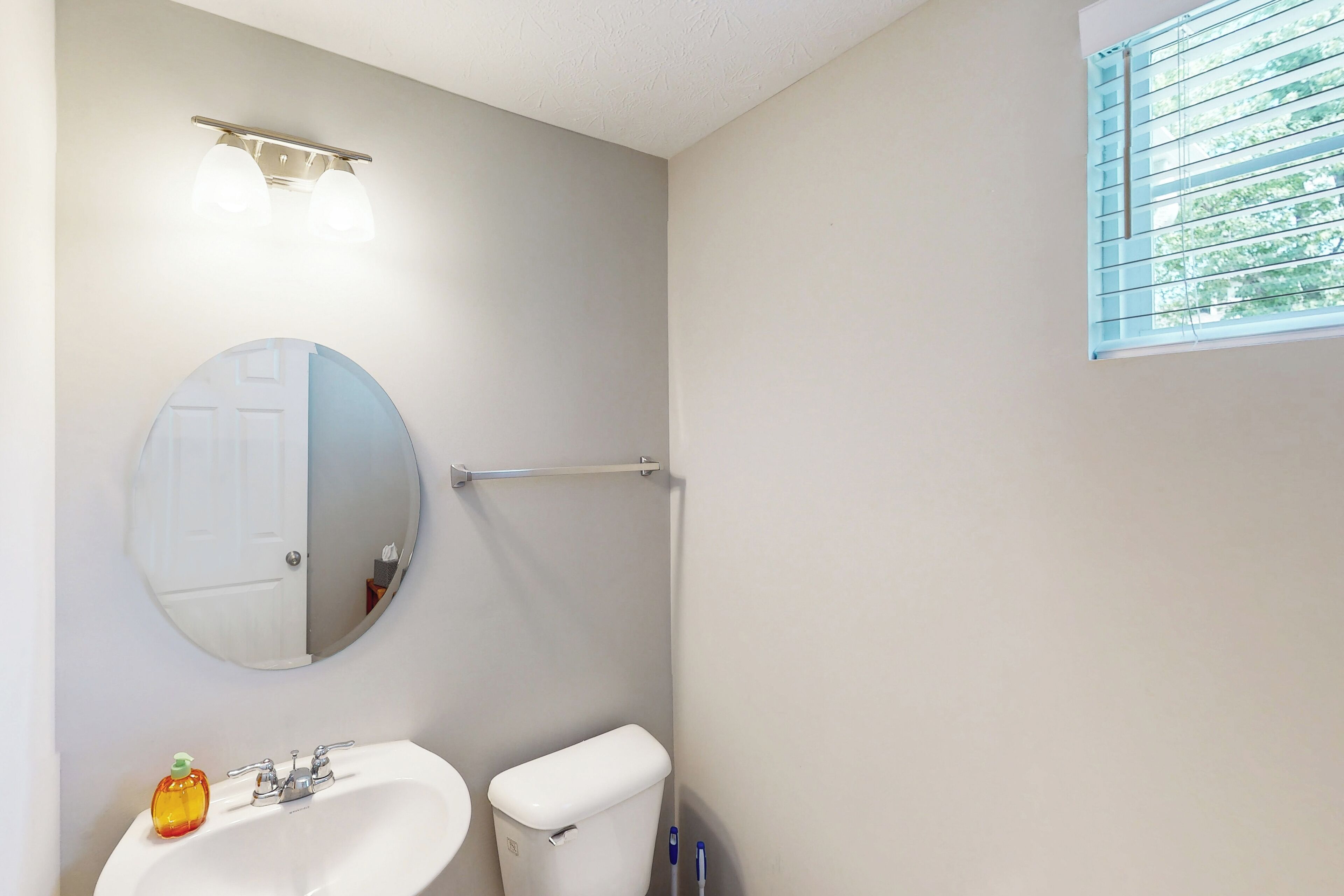 A bathroom with a round mirror, a white sink, and a toilet. There is a window with blinds and a towel rack above the sink.