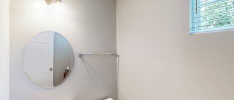 A bathroom with a round mirror, a white sink, and a toilet. There is a window with blinds and a towel rack above the sink.