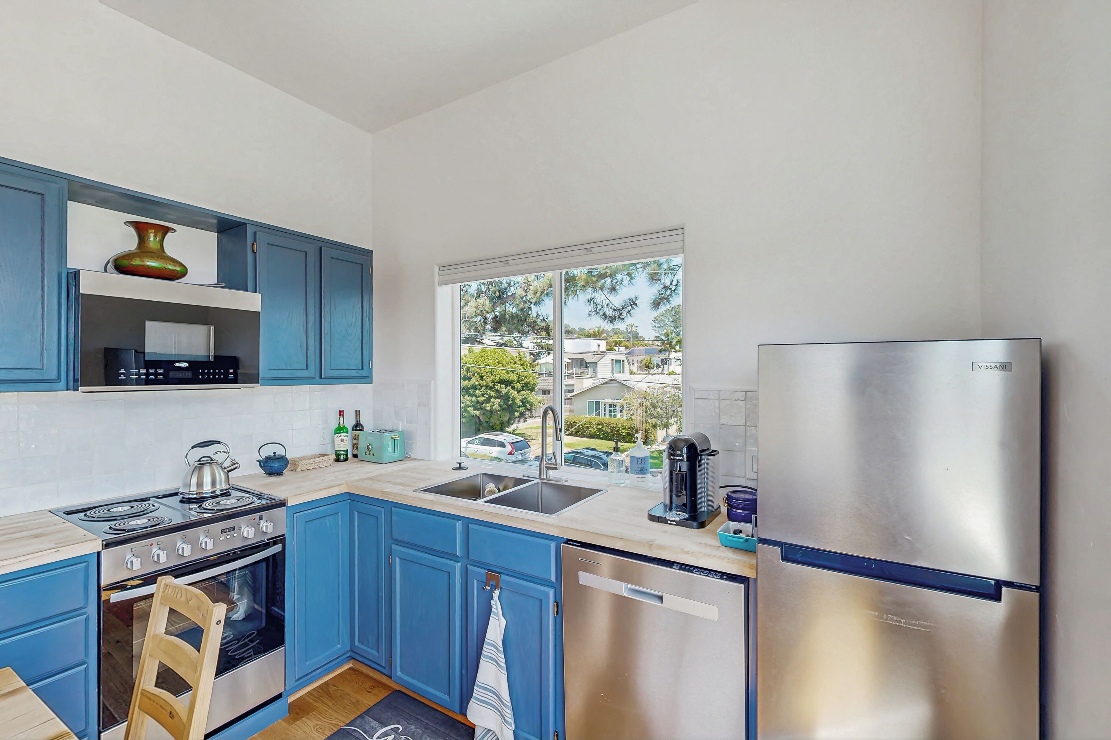 A modern kitchen with blue cabinets, stainless steel appliances, and a window offering a view of the neighborhood.