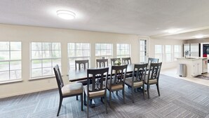A dining area with a large table and chairs, a kitchen area, and large windows with a view of trees. - 4BR w/ Pool Table & WFH Desk, Near Fishing Pond (Quinlan)