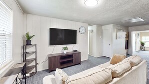 A living room with a flat-screen TV, a sofa, a coffee table, and a plant. - 4BR w/ Pool Table & WFH Desk, Near Fishing Pond (Quinlan)