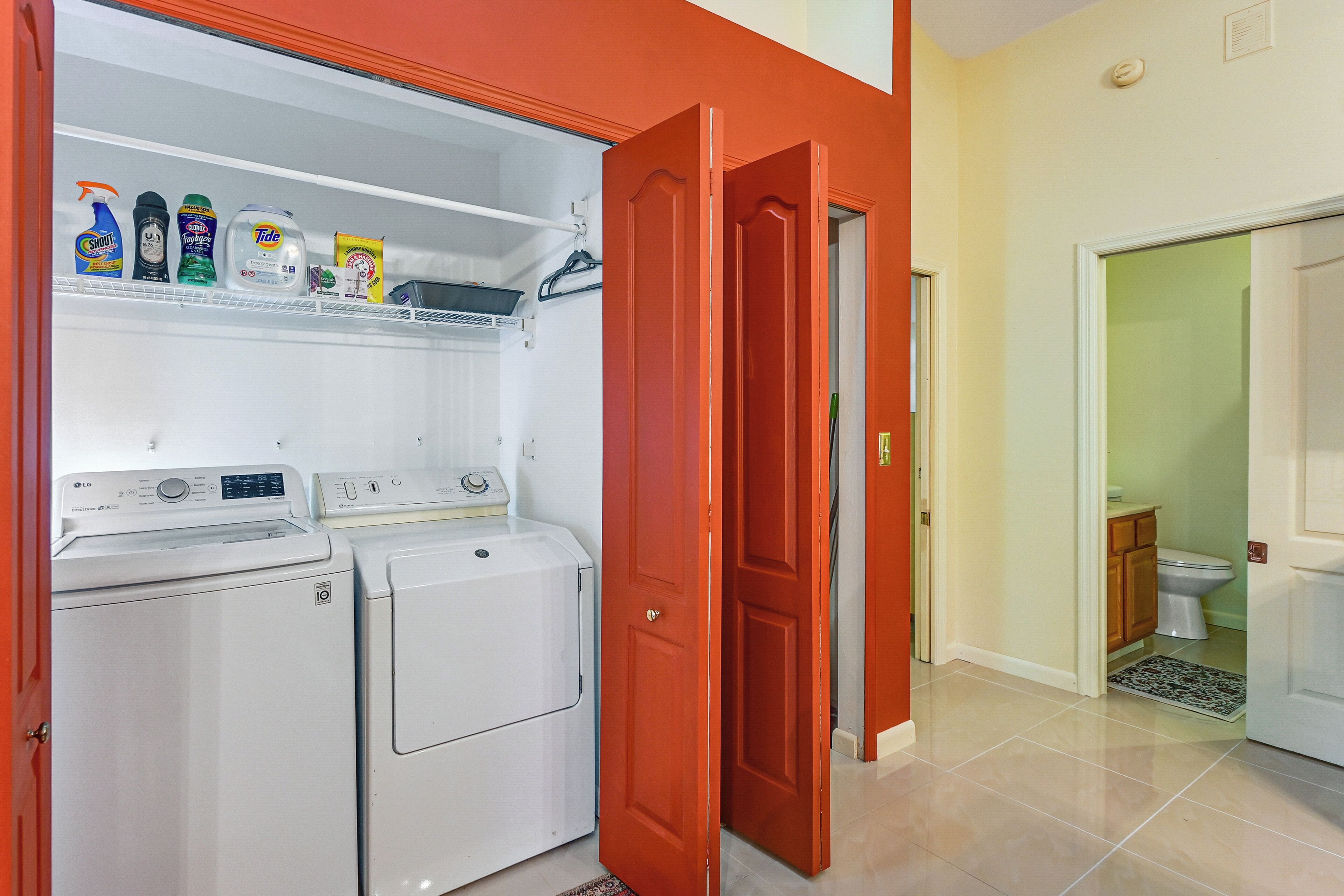A laundry room with two white washing machines, a shelf with cleaning products, and a red door leading to a bathroom.