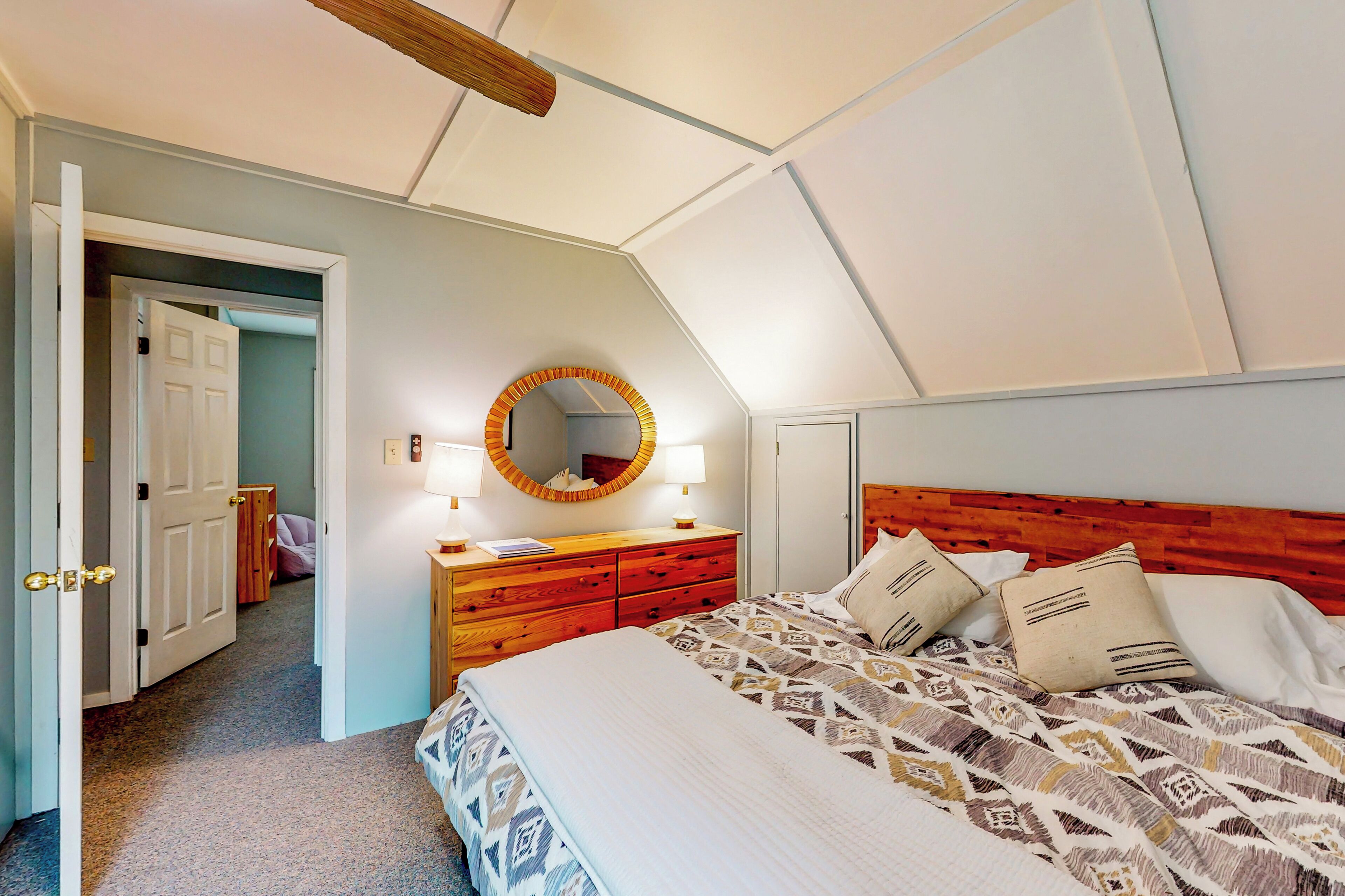 Ein Schlafzimmer mit einem Holzkopfteil, einer gemusterten Bettdecke, einem Nachttisch mit Spiegel und einer Tür zu einem anderen Raum.