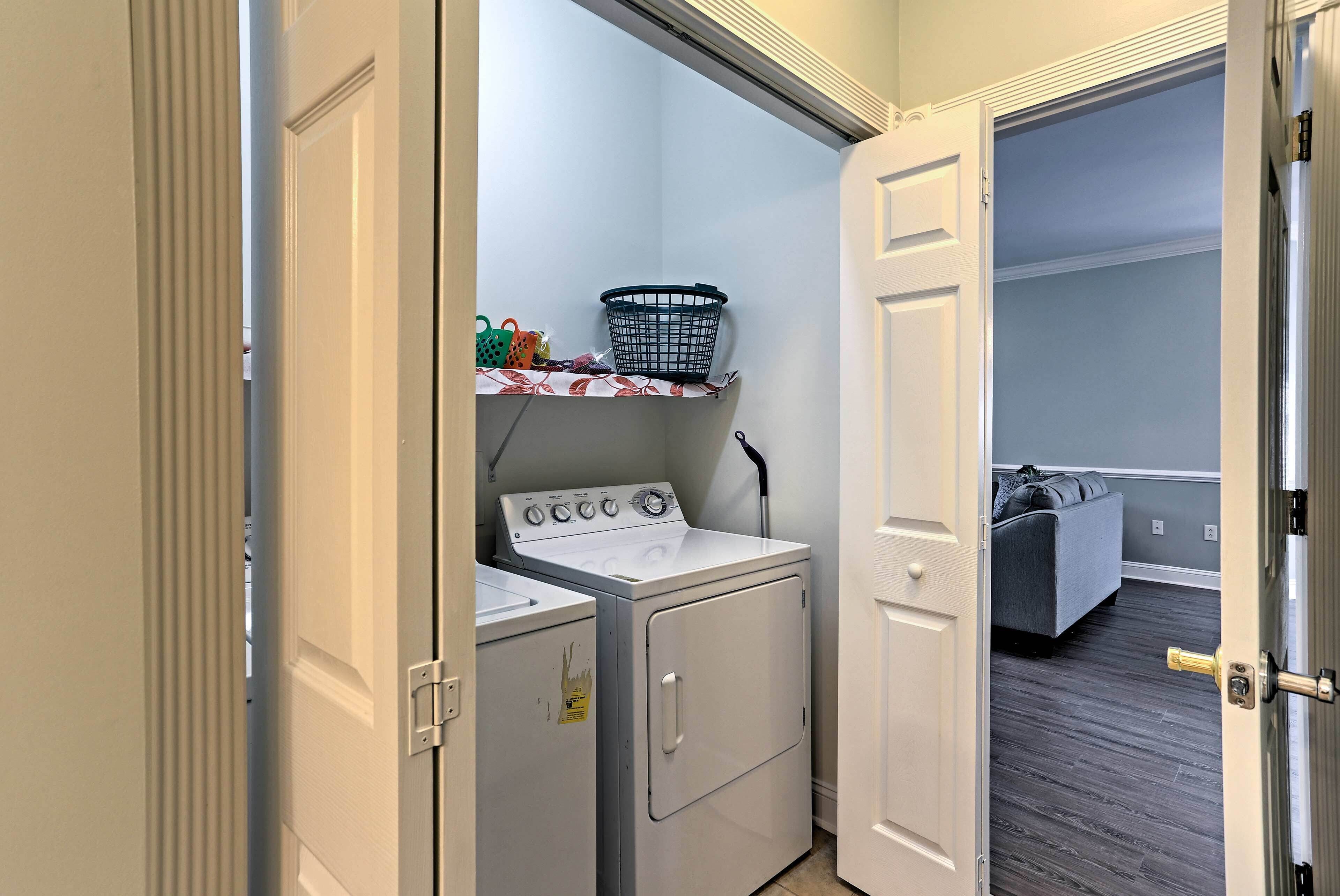 A laundry room with a washer and dryer, a shelf with laundry baskets, and a grey couch in the adjacent room.
