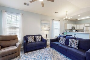 A living room with a brown armchair, two blue sofas with patterned cushions, a blue patterned rug, a ceiling fan, a chandelier, and a kitchen area with white cabinets and appliances. - 2 BR Condo w/ Shared Pool, Near Outlet Mall (Myrtle Beach)