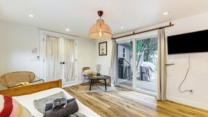 A hotel room with a wooden floor, a bed with a striped blanket, a wicker chair, a table with a lamp, and a view of the outside through a sliding door. - 5 Mi to Las Olas Beach! Bright Home w/ Fenced Yard (Fort Lauderdale)