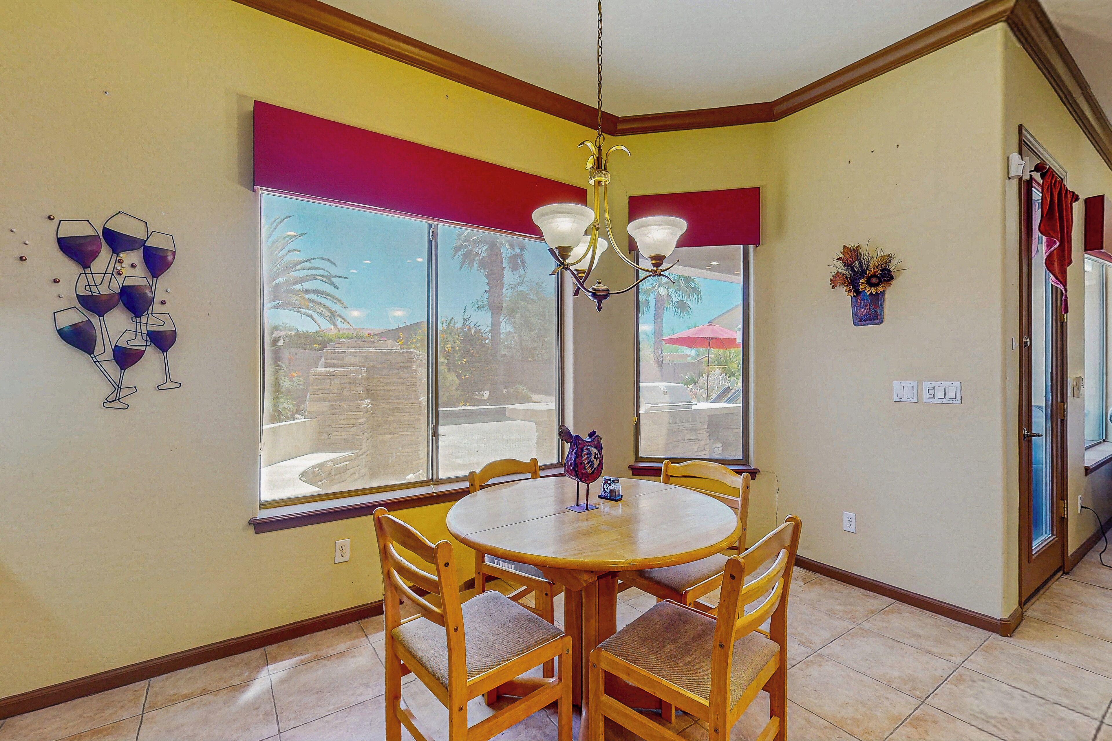 A dining area with a round table and wooden chairs, large windows with red curtains, and a chandelier.