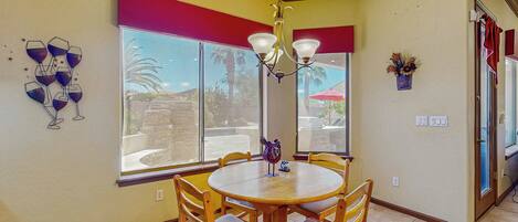 A dining area with a round table and wooden chairs, large windows with red curtains, and a chandelier.
