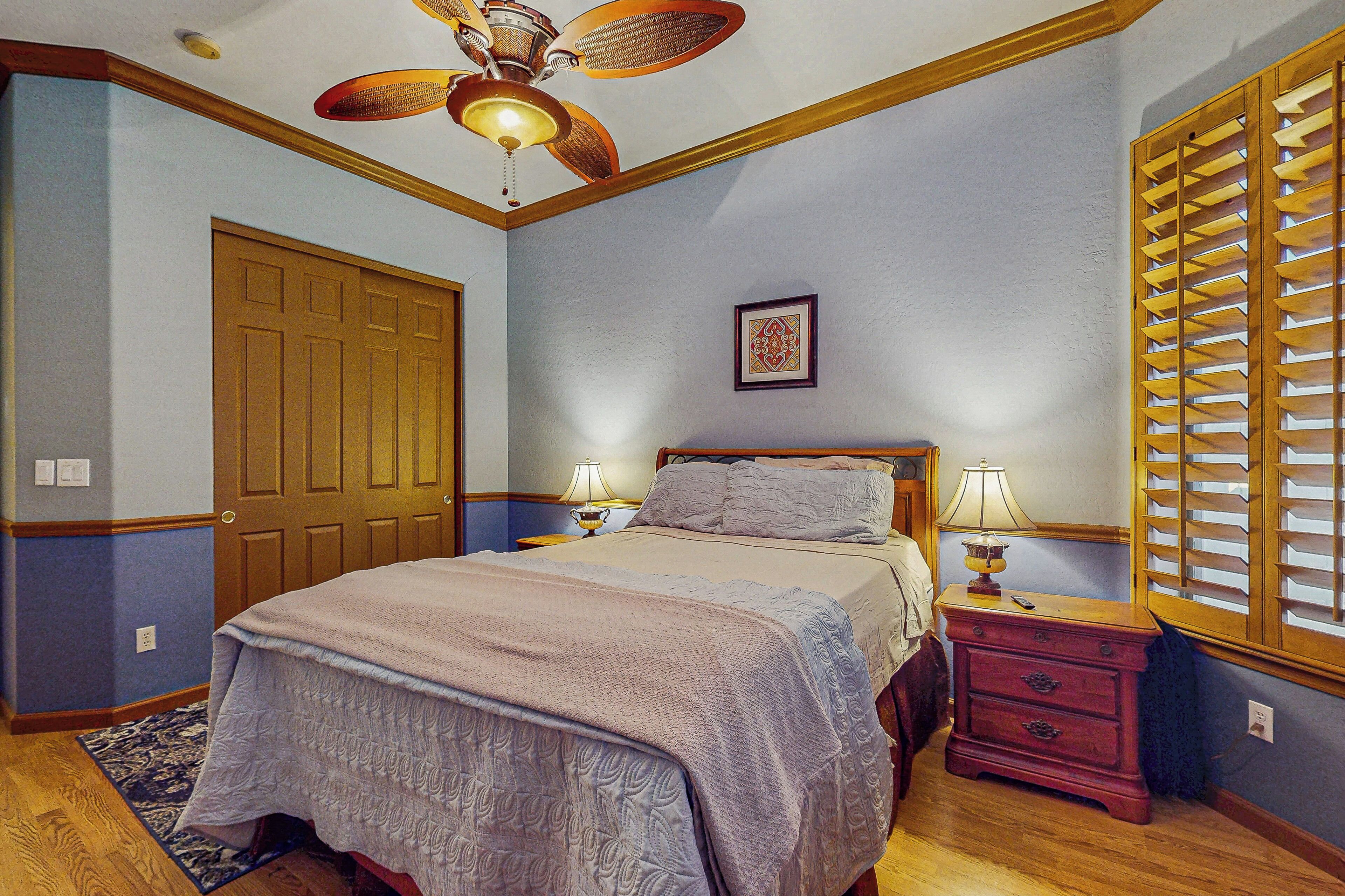 Quarto com cama, duas mesas de cabeceira com luminárias, um criado-mudo de madeira, ventilador de teto, porta e janela com persianas.