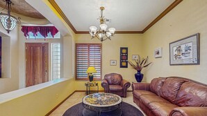 A living room with a brown leather sofa, a round coffee table with a patterned top, a chandelier, and a window with blinds. - Goodyear Home w/ Private Pool, Spa & Built-in BBQ (Goodyear)