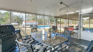Patio esterno con tavolo in vetro, sedie in metallo, barbecue e vista sulla piscina e sul giardino.