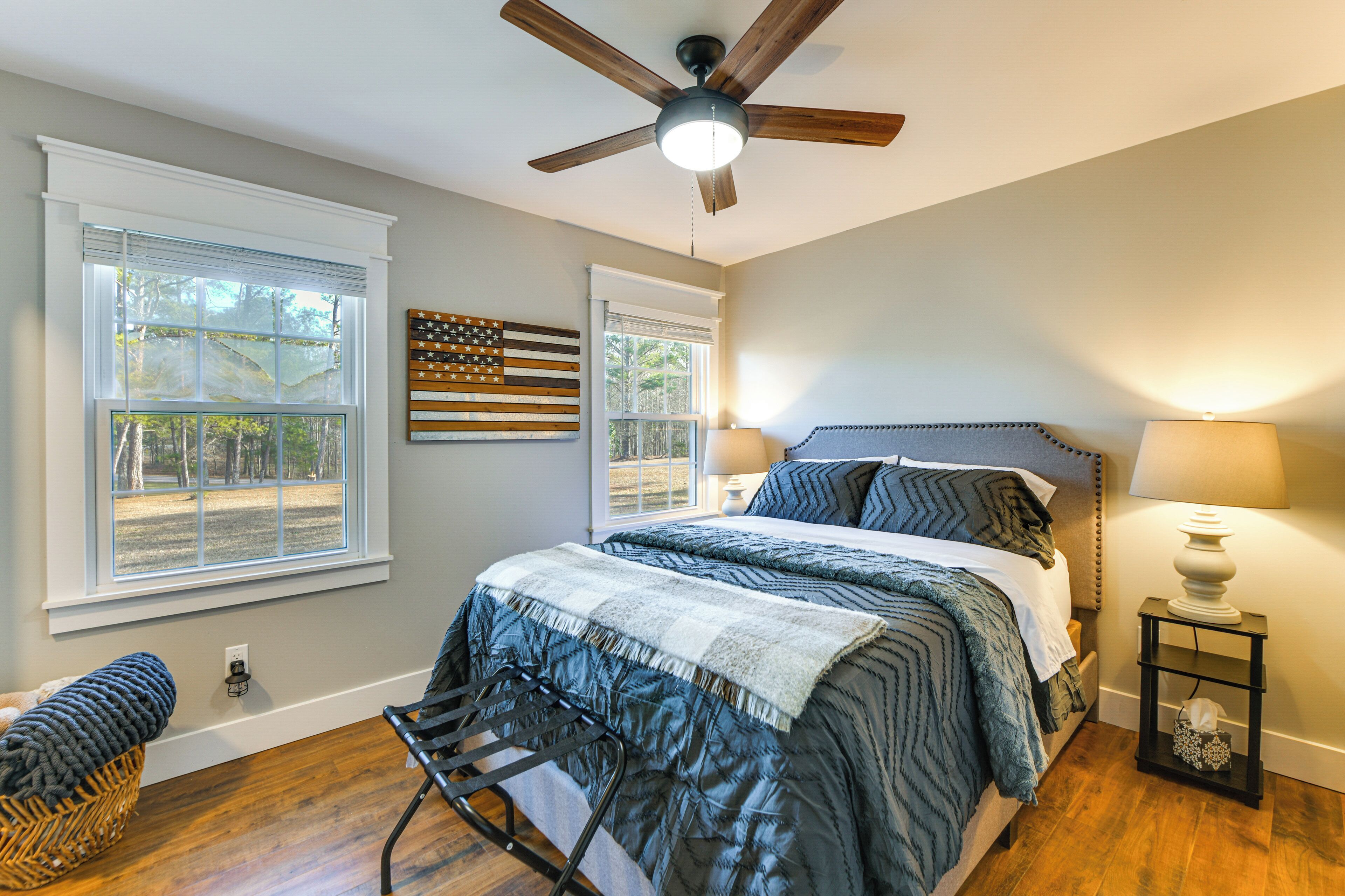 A bedroom with a bed, two windows, a ceiling fan, and a nightstand with a lamp.