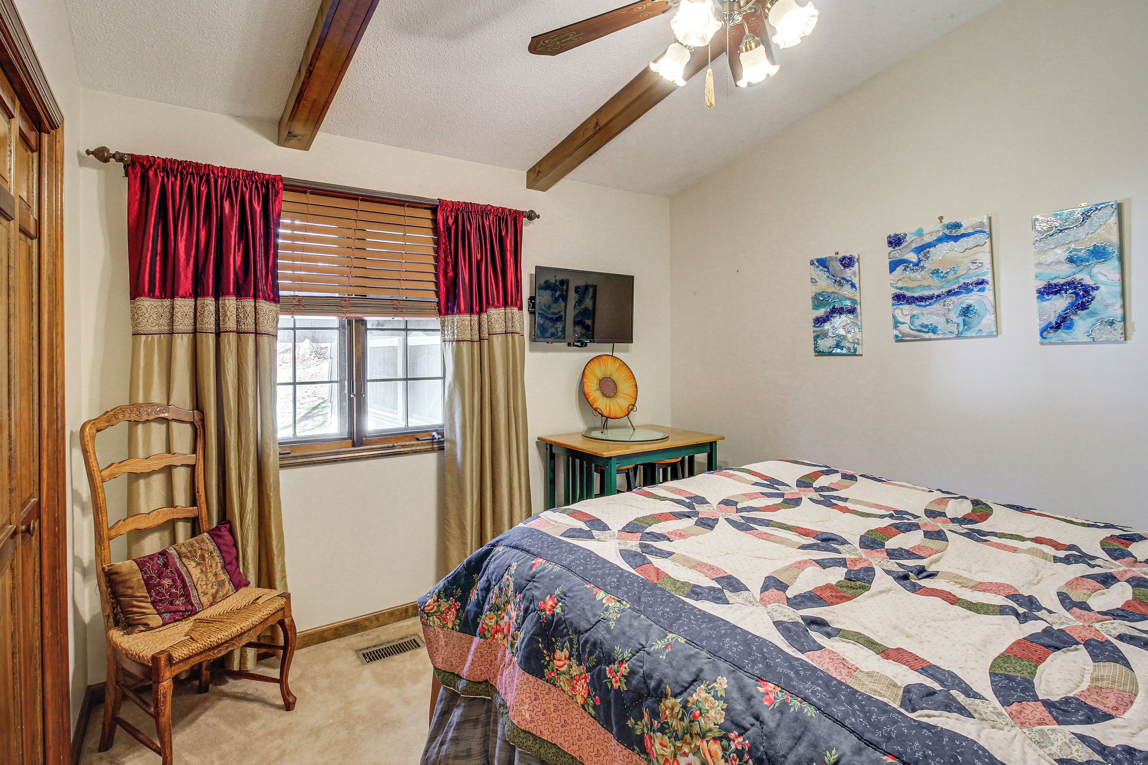 Une chambre avec un grand lit, une chaise en bois, une fenêtre avec des rideaux, un ventilateur de plafond et des œuvres d’art murales.