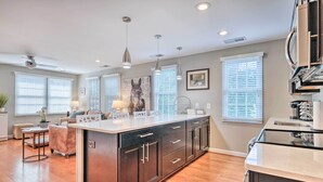 A modern kitchen with a central island, pendant lighting, and a large window with blinds. - Old Town Walkable Paradise w/ Private Patio! (Alexandria)