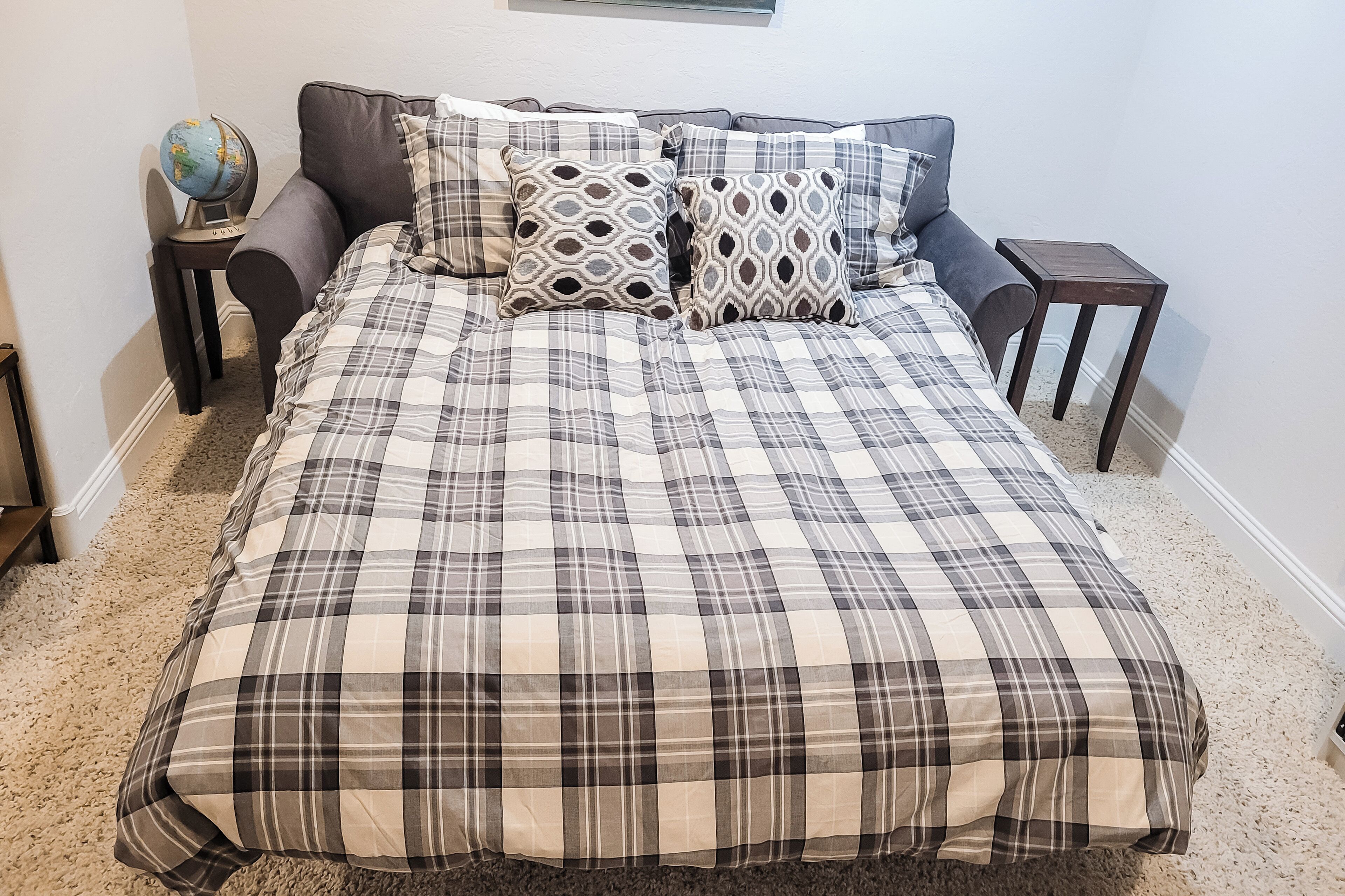 Een bed met een geruite sprei, twee gestreepte kussens, een wereldbol op een bijzettafeltje en een klein houten nachtkastje.
