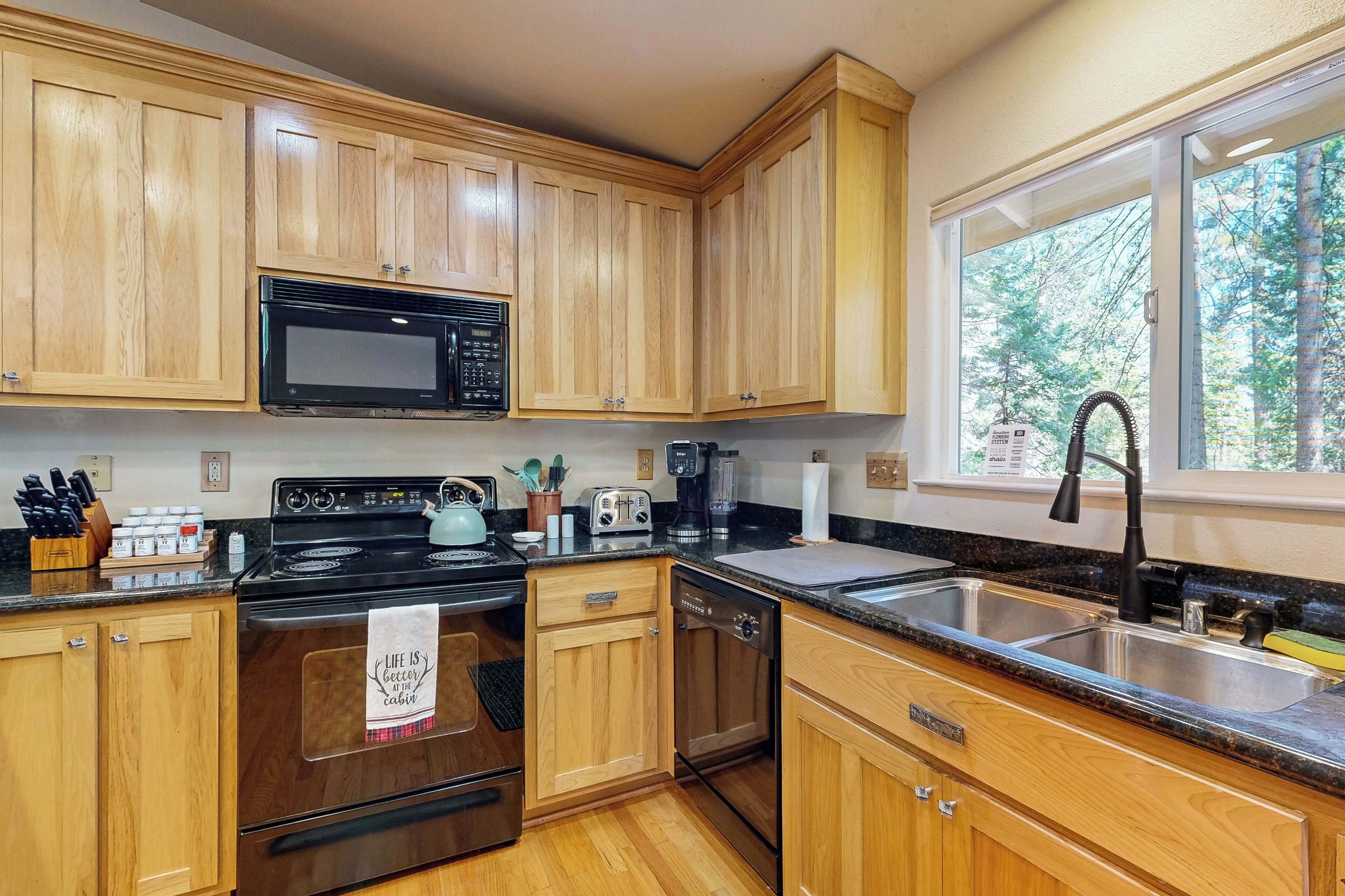 A modern kitchen with wooden cabinets, a black microwave, stainless steel appliances, and a large window offering a view of trees.