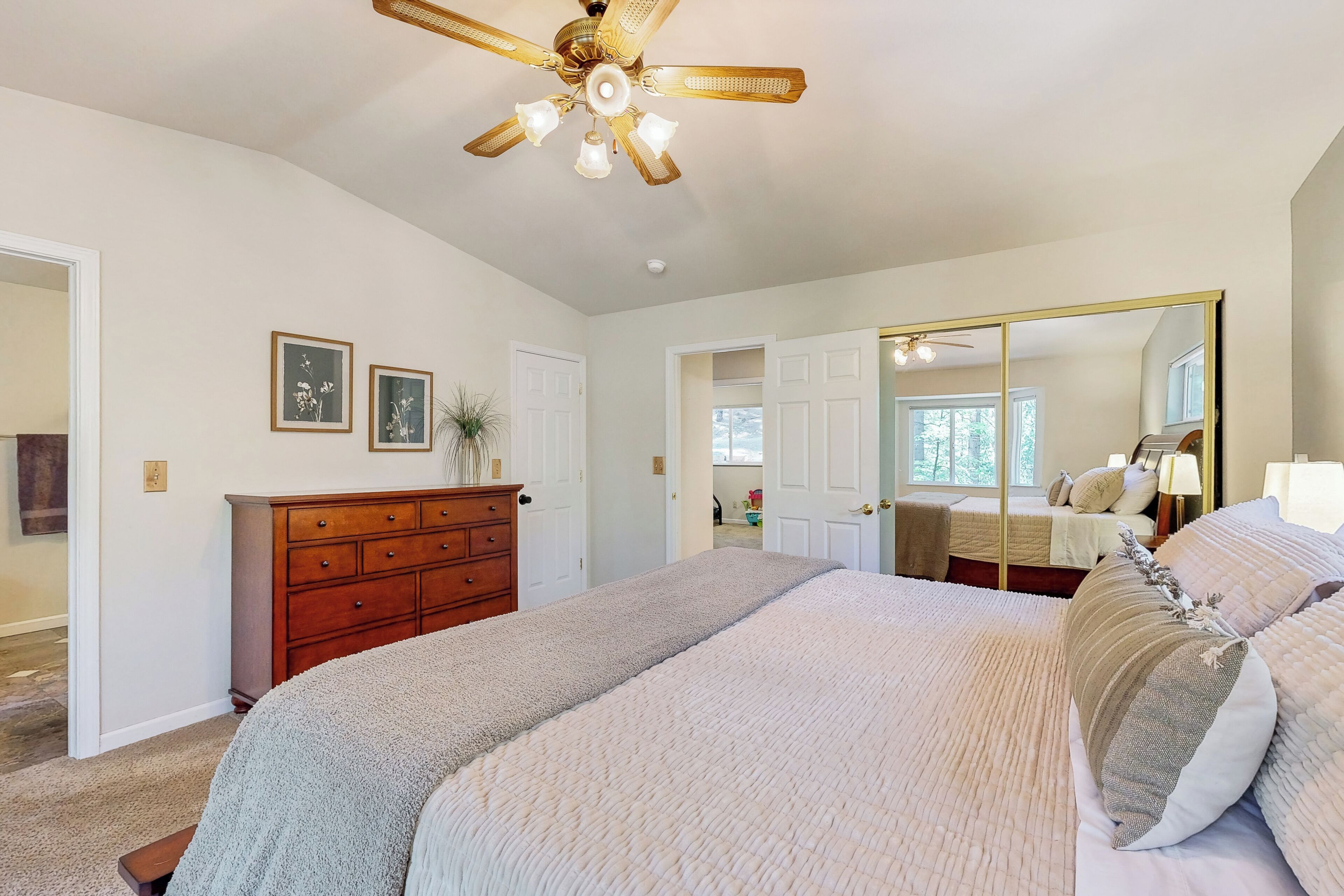 Une chambre avec un grand lit, une commode en bois, un ventilateur de plafond et un grand miroir.