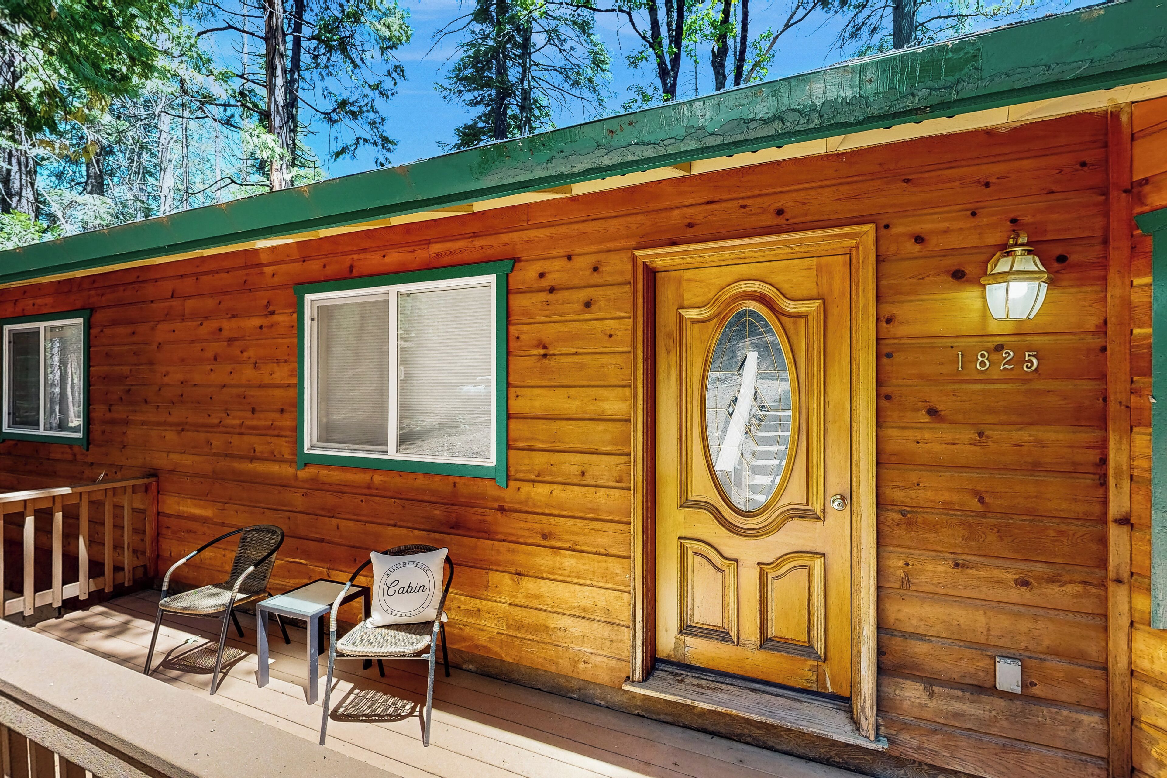 A wooden cabin with a large ornate door, a small porch with two chairs, and a window with a green frame.