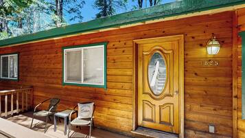 A wooden cabin with a large ornate door, a small porch with two chairs, and a window with a green frame.