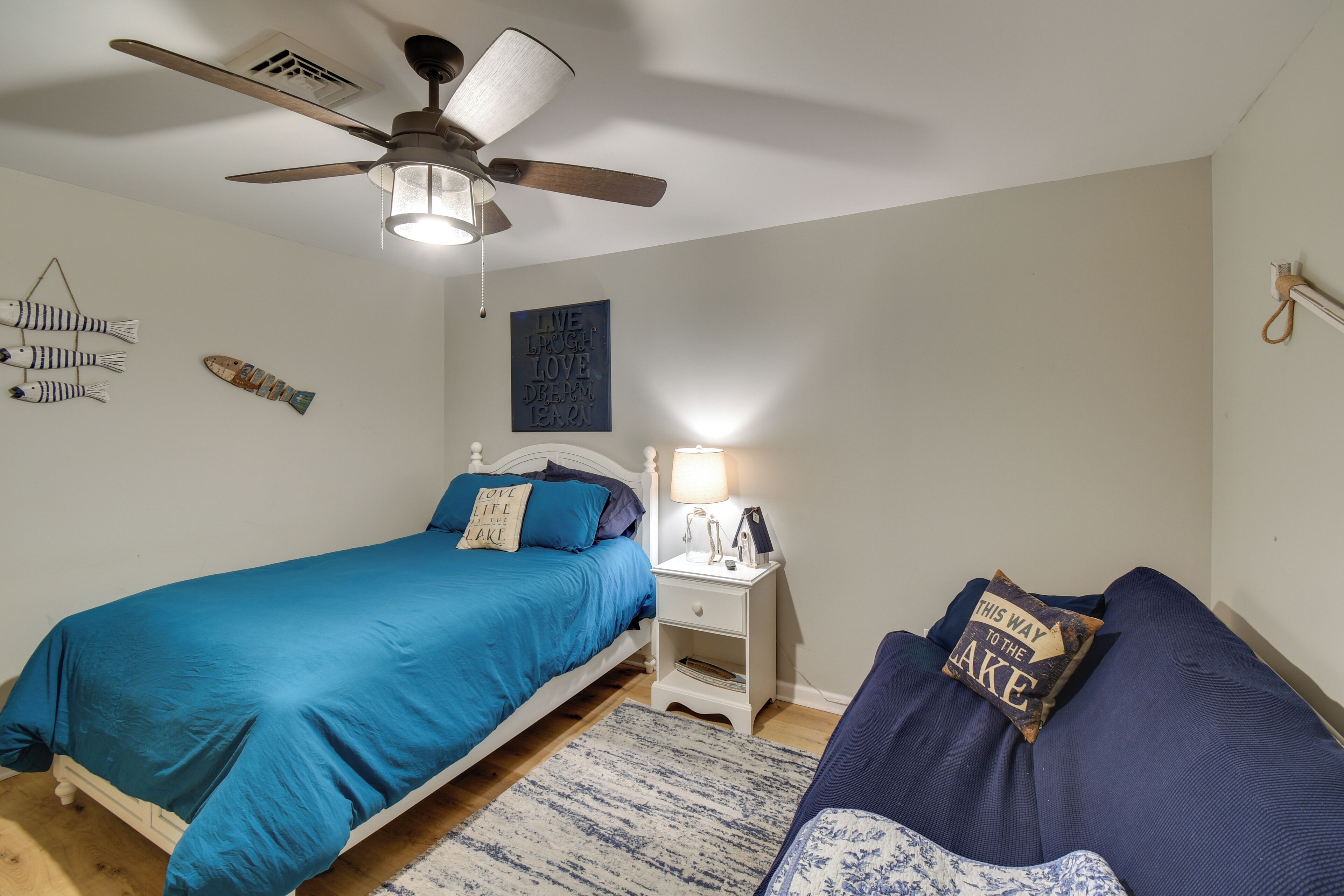 A bedroom with two beds, a ceiling fan, a nightstand with a lamp, and wall decorations.