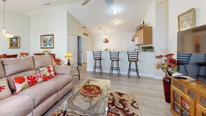 A living room with a sofa, coffee table, and a TV. There is a kitchen area with a bar and chairs. - Dog-friendly Home w/ Golf Cart, AC, Wifi (The Villages)
