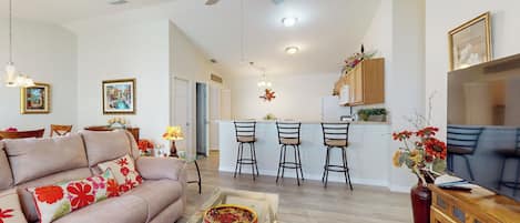 A living room with a sofa, coffee table, and a TV. There is a kitchen area with a bar and chairs.