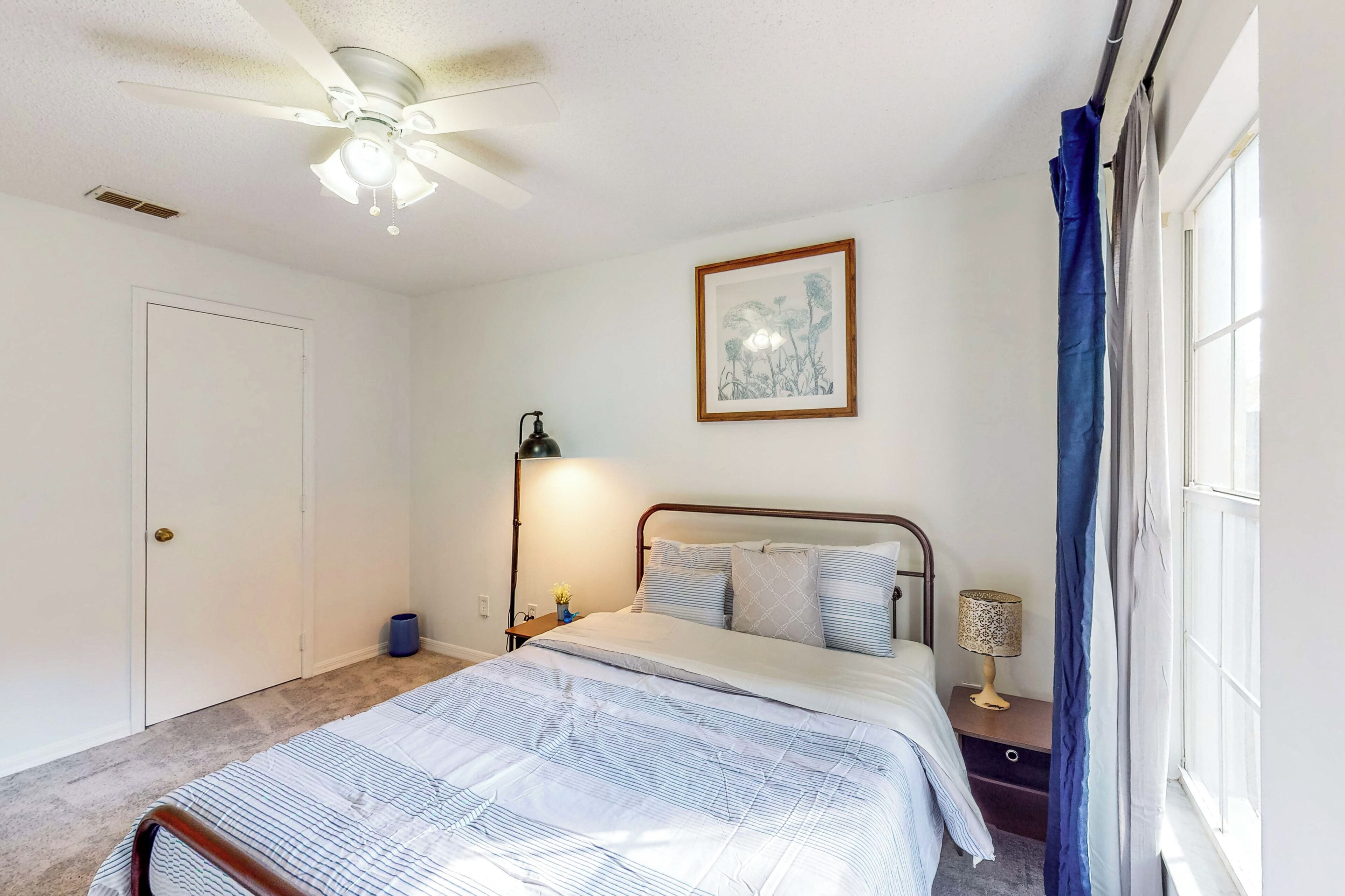 A bedroom with a bed, a ceiling fan, a nightstand with a lamp, a framed picture on the wall, and a window with curtains.