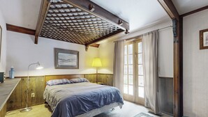 Un dormitorio con cama, escritorio, lámpara, ventana con cortinas y estructura de techo de madera.