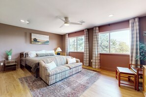 A bedroom with a bed, a sofa, a wooden chair, a ceiling fan, and a large window with curtains. - Buckthorn Beauty: Golf, Hike & More (Show Low)