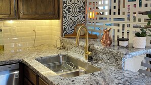 Una cucina con piani di lavoro in granito, lavello in acciaio inox, mobili in legno e un decorativo boccale di rame.